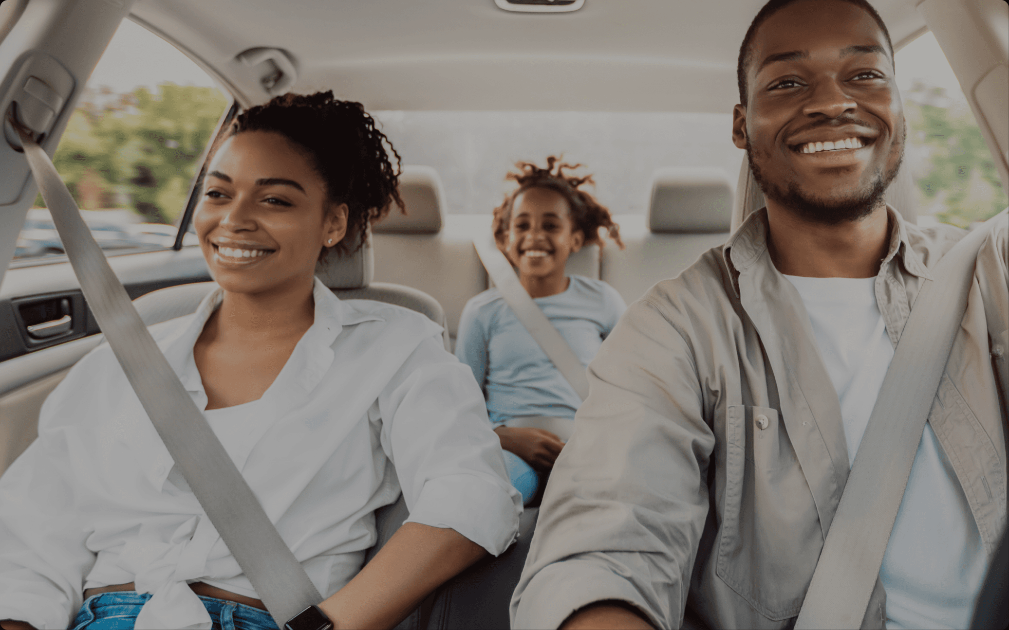 A family of three smiling in a car, all wearing seatbelts